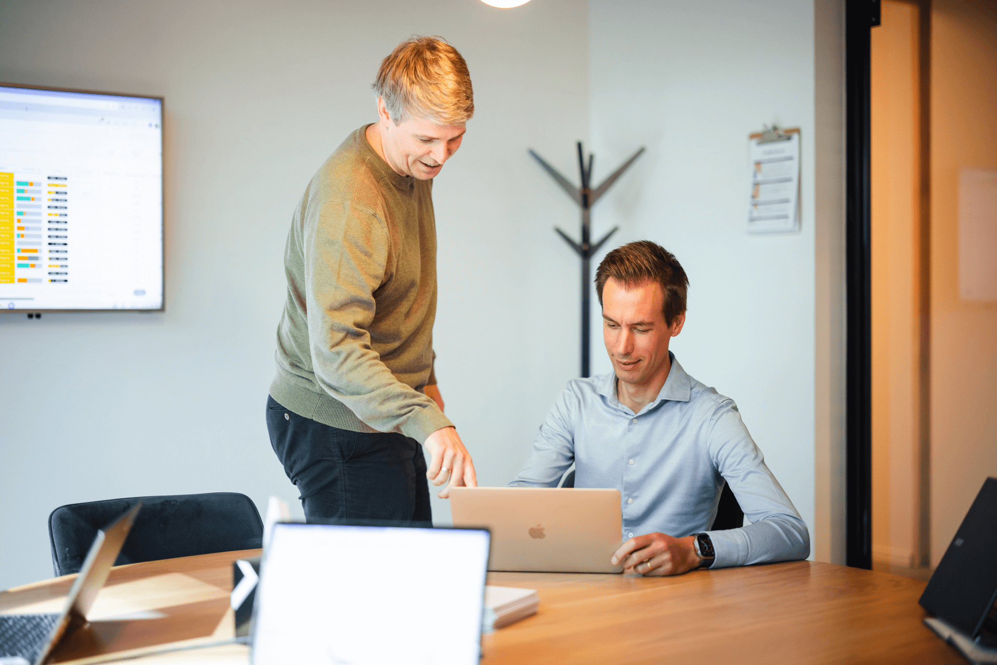 Two colleagues collaborating in a bright office; one is seated at a desk while the other leans over to explain something on the screen during monday.com training.